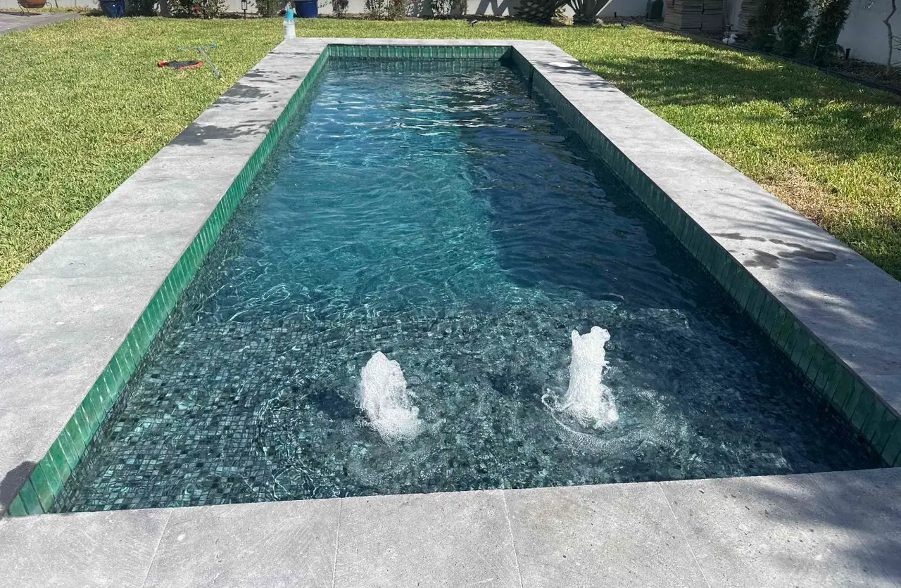 Long lap pool with green glass waterline tile and two bubblers running on a bright Laredo afternoon