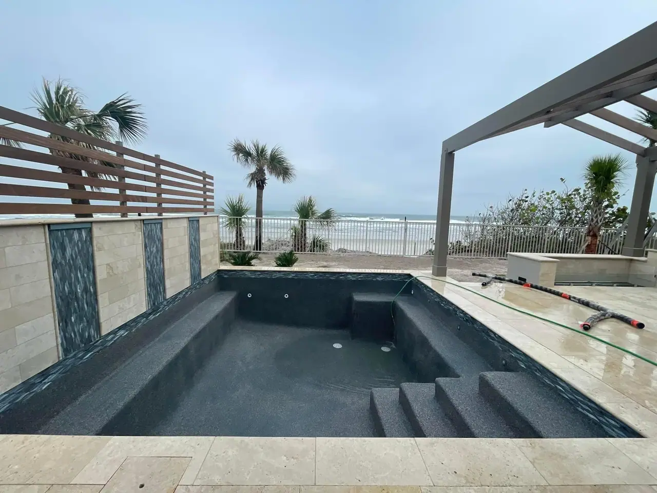 Newly plastered dark pool and spa with travertine coping and blue mosaic tile, a pergola and palms, and the ocean beyond the fence