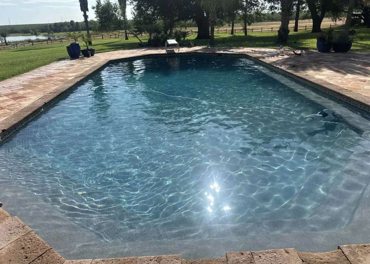 Finished rectangular pool with clear water and a light stone deck, a lounge chair and potted plants at the far end, and a fenced pasture, pond and trees behind
