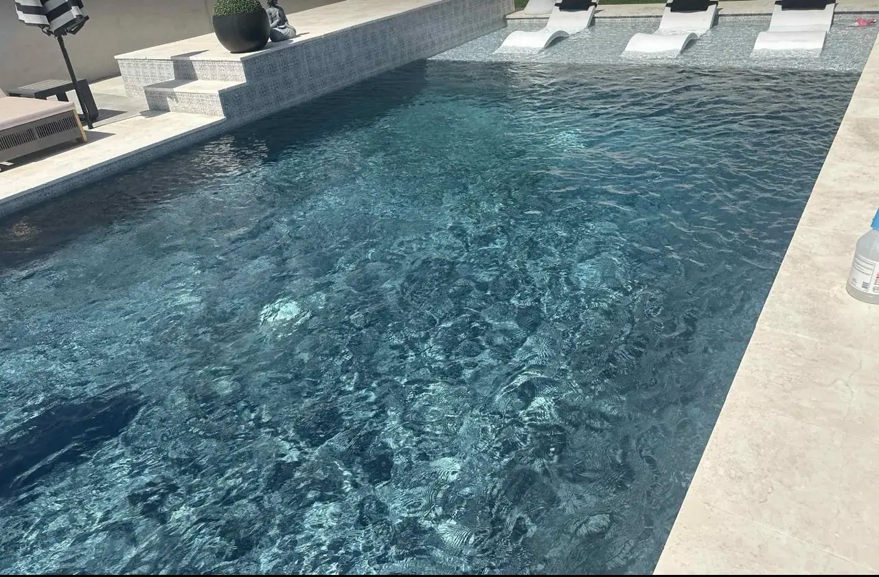 Dark water in a rectangular pool with a mosaic tiled step, travertine coping and white in-water loungers along the far ledge