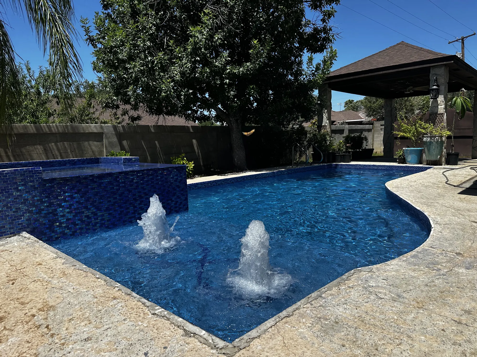 Two water jets rising out of a deep blue tiled pool, with a stone deck, a palm and a gazebo behind it