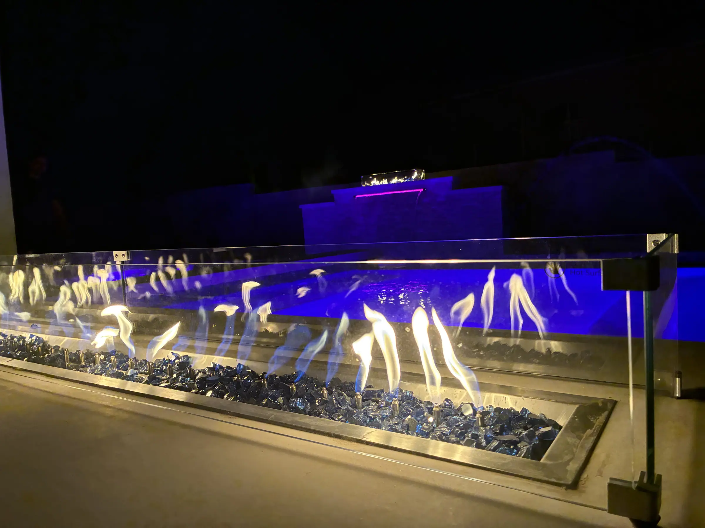 Linear fire feature burning beside a blue-lit JCW pool wall after dark
