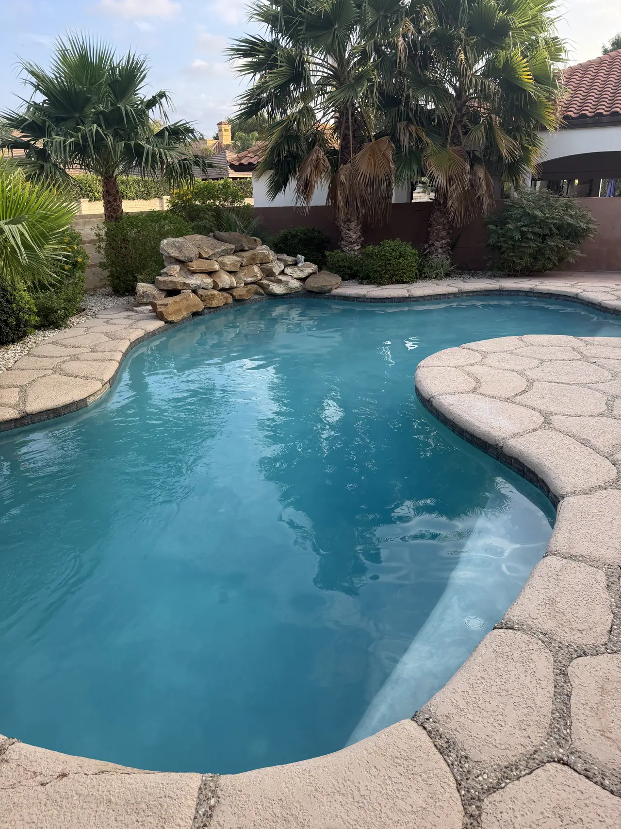 Freeform JCW pool with a natural stone deck and palms beside a tile-roofed Laredo home