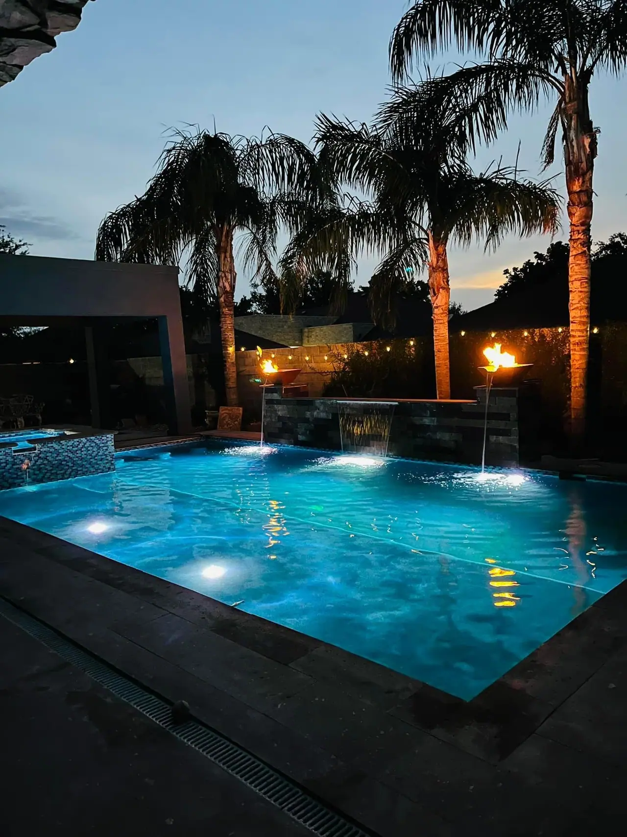 JCW pool lit at twilight with fire bowls burning on the raised wall and palms overhead