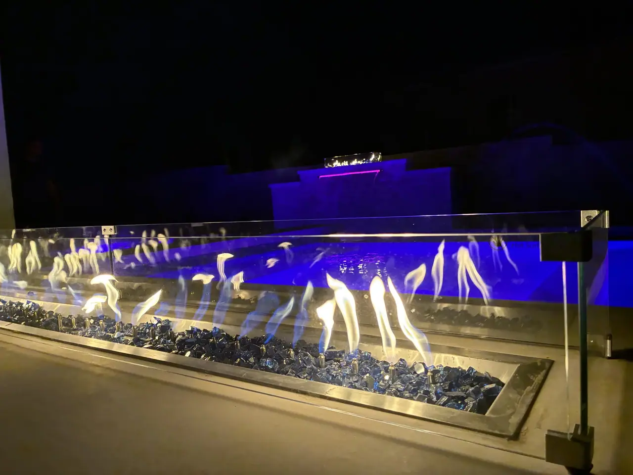 Linear fire feature burning over blue fire glass behind a glass guard at night, with a blue lit pool and a spillway wall behind it