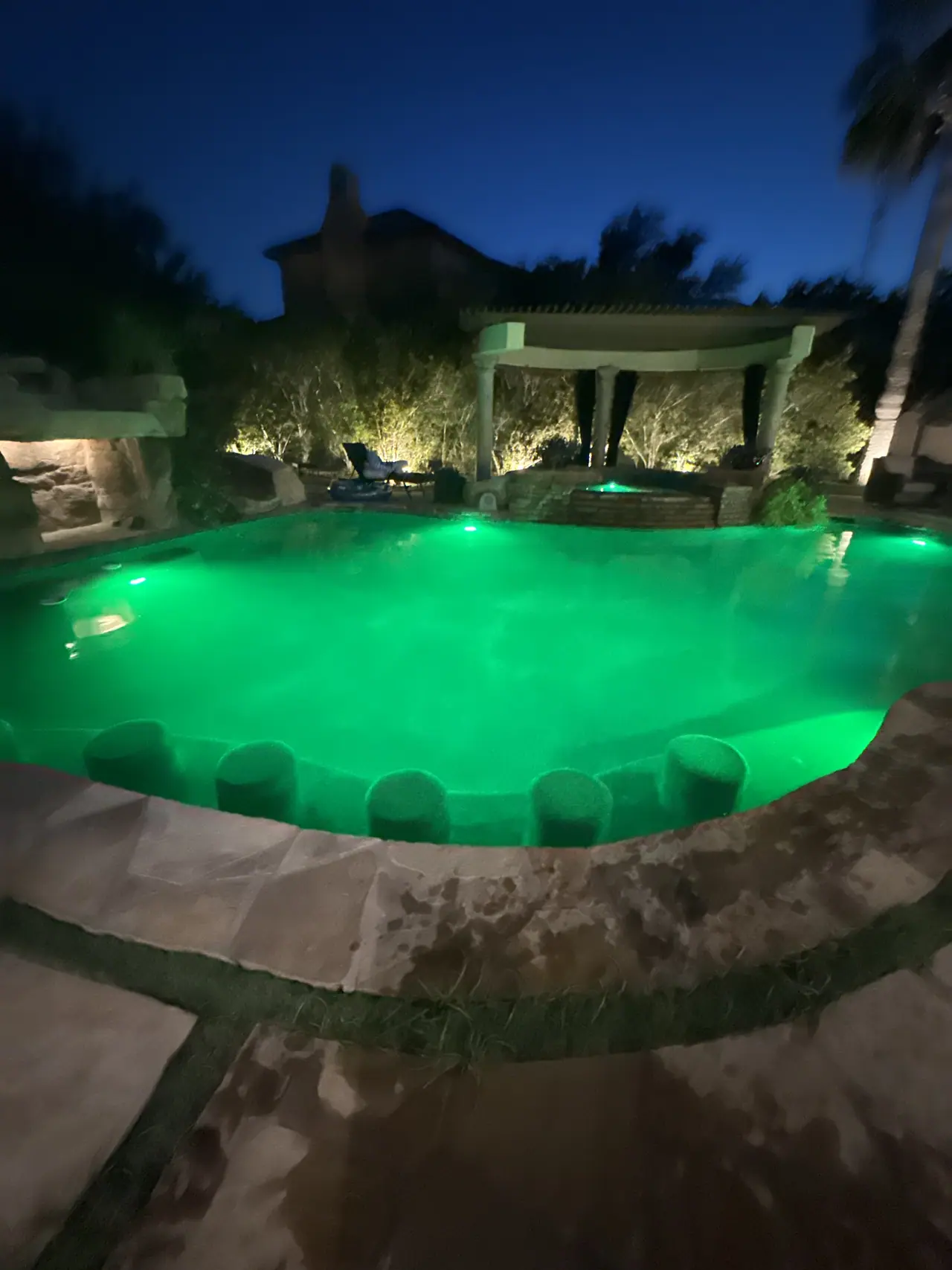 Freeform JCW pool lit green after dark with an illuminated pergola behind it
