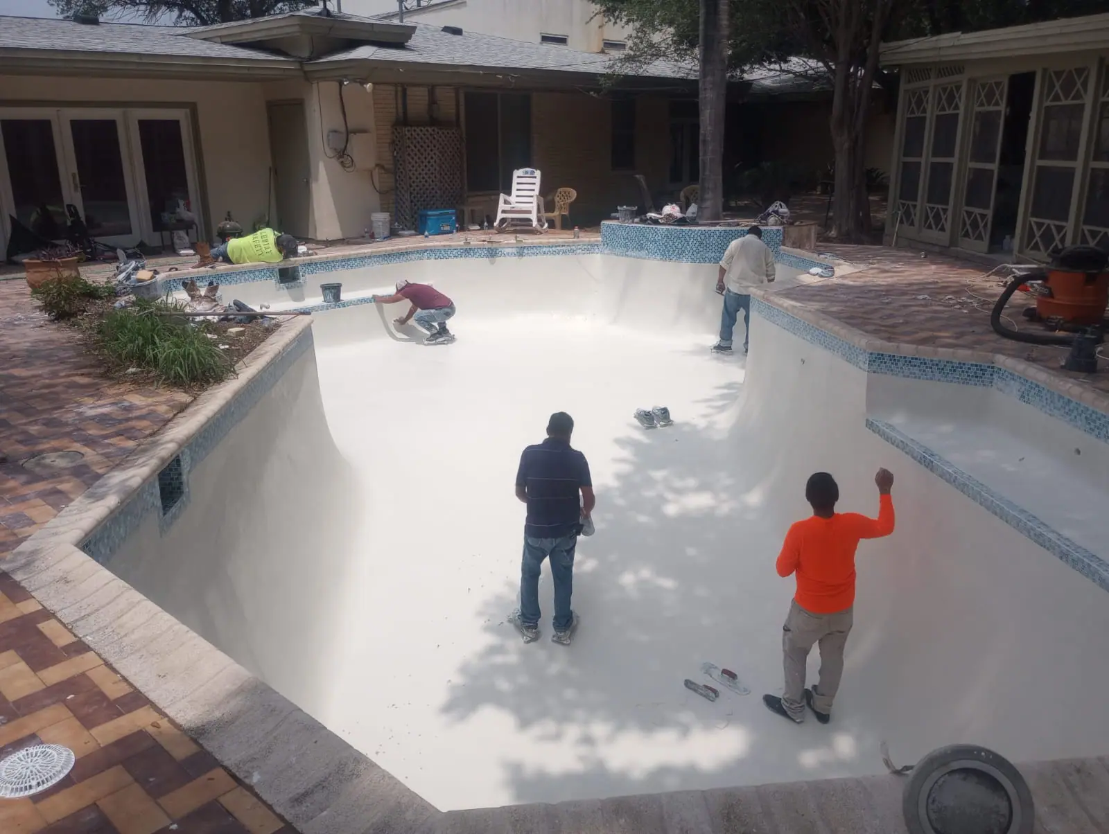 Three-person JCW crew finishing new white plaster across a drained pool shell beside a brick deck